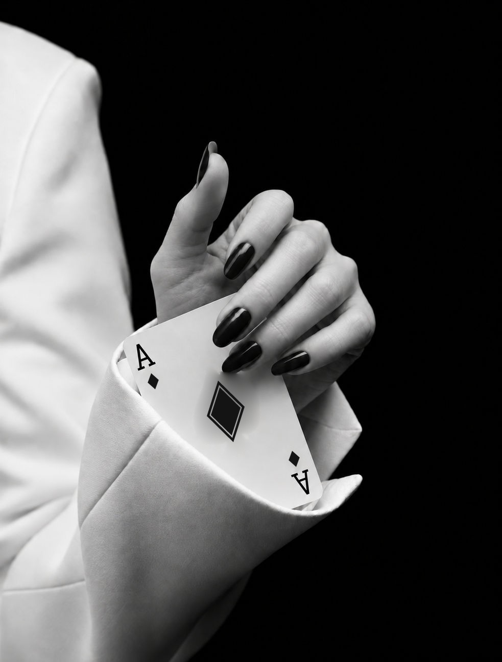 Mão de unhas escuras tirando um ás de ouros da manga de um terno branco, em preto e branco